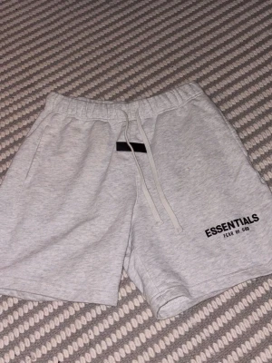 Grå Essentials Fear of God shorts - Snygga ljusgrå shorts från Essentials Fear of God med snörning i midjan och tryckt logga på vänster ben. Tillverkade i mjuk bomull för maximal komfort. Perfekta för chill dagar eller när du vill ha en clean look.