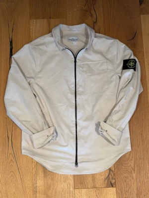 Vit overshirt från Stone Island - Säljer en stilren vit overshirt från Stone Island med svart dragkedja framtill och klassisk patch på vänster ärm. Skjortan har en bröstficka och är tillverkad i ett lätt, mjukt material som passar perfekt för lager-på-lager. Snygg och enkel att matcha till olika outfits.