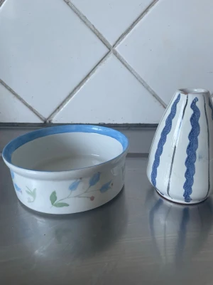 Antika gamla saker  Keramikskål och liten vas - Antika gamla sakerna Säljer en rund keramikskål med blå kant och blommönster samt en liten vit vas med blå vågiga ränder. Perfekt för dekoration eller småsaker. Båda är i keramik och har en stilren design som passar i de flesta hem.