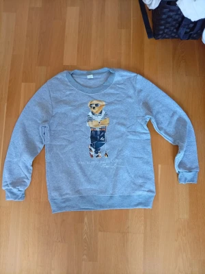 Grå sweatshirt med Polo Bear-tryck - Snygg grå sweatshirt från Ralph Lauren med det ikoniska Polo Bear-trycket framtill. Rund halsringning och ribbade muddar vid ärmslut och nederkant. Perfekt för en avslappnad och trendig look.