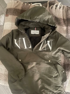 Olivgrön vindjacka från Valentino - Snygg olivgrön vindjacka från Valentino med stor vit VLTN-logga på bröstet. Jackan har huva, magficka med knappar och svart innerfoder. Perfekt för dig som vill ha en streetig och trendig look.