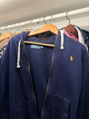 Marinblå hoodie från Polo Ralph Lauren - Mysig marinblå hoodie från Polo Ralph Lauren med dragkedja, två stora fickor framtill och vit dragsko i huvan. Klassisk gul logga broderad på bröstet. Tillverkad i mjukt bomullsmaterial som är perfekt för chill dagar.