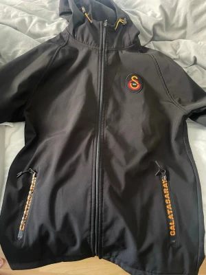 Svart vindjacka Galatasaray - Svart vindjacka med huva och dragkedja framtill. Jackan har Galatasaray-logga på bröstet och gula detaljer på snören och dragkedjor. Två fickor med dragkedja och Galatasaray-tryck. Perfekt för dig som vill visa ditt stöd för laget.
