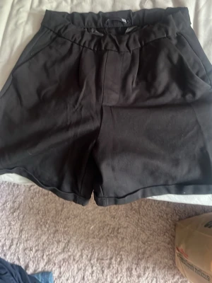 Svarta högmidjade shorts - Säljer ett par svarta högmidjade shorts med avslappnad passform och resår i midjan. Shortsen har fickor fram och dekorfickor bak. Perfekta till sommaren och lätta att matcha med olika stilar.