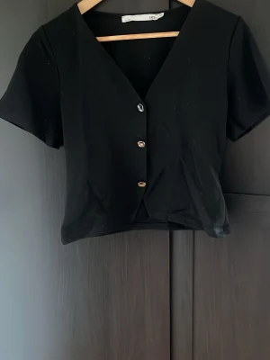 Svart kortärmad topp med knappar - Stilren svart topp från lager 157 med v-ringning och silvriga knappar. Kortärmad och croppad modell som passar perfekt till både jeans och kjol. Använd ett fåtal gånger och är som ny.