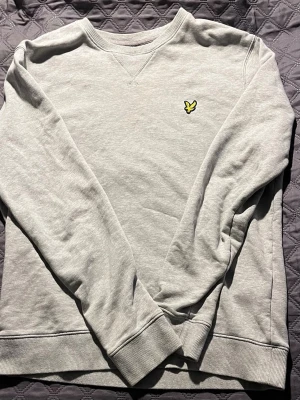 Ljusgrå lyle&scott sweatshirt - Ljusgrå sweatshirt med rund hals och ribbade muddar. På bröstet sitter en liten broderad gul logga i form av en fågel. Tröjan har en avslappnad passform och är tillverkad i mjukt bomullsmaterial. Perfekt för en chill och stilren look.