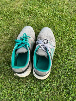 Grå sneakers med turkosa detaljer - Säljer ett par grå sneakers med turkosa och svarta detaljer samt meshmaterial för bra ventilation. Skorna har olika färg på skosnörena, ett turkost och ett grått, och insidan är fodrad i turkost tyg. Klassisk låg modell med vit sula.