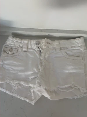 Vita fransiga jeansshorts - Säljer ett par vita jeansshorts med fransig kant och klassisk femficksdesign. Shortsen har bälteshällor, knapp- och dragkedjestängning samt dekorativa sömmar. Perfekta för varma dagar och enkel att matcha med olika toppar. Storlek 146 