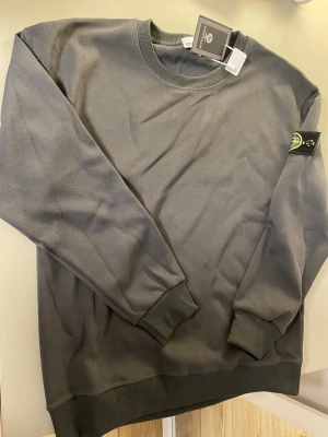 Mörkgrå sweatshirt med logotypärm - Snygg mörkgrå sweatshirt med rund hals och ribbade muddar vid ärmslut och nederkant. Tröjan har en tydlig logotyp-patch på vänster ärm och är tillverkad i mjukt bomullsmaterial. Perfekt för en avslappnad och trendig stil.