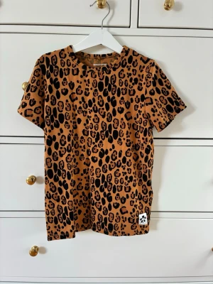 Leopardmönstrad t-shirt Mini Rodini - T-shirt från Mini Rodini med leopardmönster i svart på brun botten. Klassisk rund halsringning och korta ärmar. Strl 128/134. Fint skick används fåtal gånger.