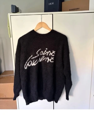 Svart stickad Saint Laurent-tröja - Snygg svart stickad tröja från Saint Laurent med vit logga i handskriven stil på bröstet. Tröjan har rund halsringning, långa ärmar och ribbade muddar. Perfekt för dig som vill ha en trendig och bekväm look.