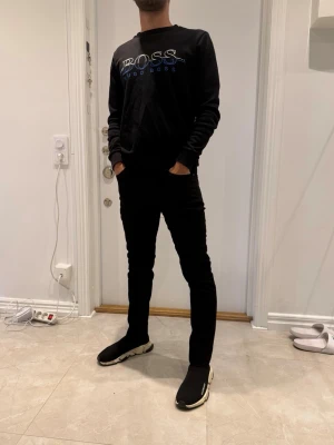 Svart hoodie från BOSS - Svart hoodie från BOSS med tryckt logga i vitt och blått på bröstet. Klassisk modell med långa ärmar och ribbade muddar. Perfekt för en stilren och avslappnad look.