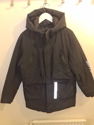 Svart pufferjacka från adidas Terrex - Säljer en svart pufferjacka från adidas Terrex med stor huva, två dragkedjeförsedda bröstfickor och reflekterande detaljer. Jackan har adidas Terrex-logga i vitt på ena ärmen och är perfekt för kalla vinterdagar. Materialet är slitstarkt och vattenavvisande.