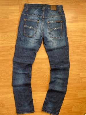 Nudie Jeans Thin finn modell  - Säljer ett par klassiska blå jeans från Nudie Jeans med snygg slitning och kontrastsömmar. Jeansen har raka ben, två bakfickor med vågig brodyr och läderpatch i midjan. Perfekta för en avslappnad och trendig look.