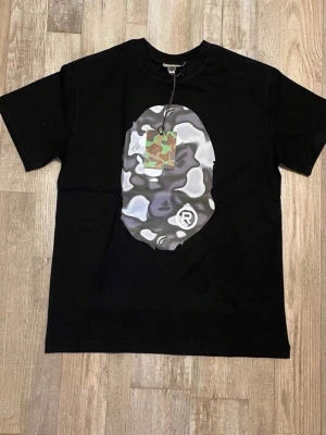 Svart t-shirt med stor ape-tryck - Säljer en svart t-shirt med ett stort, grått och vitt ape-tryck på bröstet. T-shirten har rund hals och korta ärmar. Trycket har en cool, nästan camo-liknande effekt som gör den unik och sticker ut.