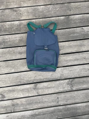 Blå och grön ryggsäck från Lacoste - Snygg blå ryggsäck från Lacoste med gröna detaljer och gröna axelband. Ryggsäcken har ett stort huvudfack med lock och spänne samt en framficka med präglad Lacoste-logga. Tillverkad i slitstarkt tyg, perfekt för skolan eller utflykter.