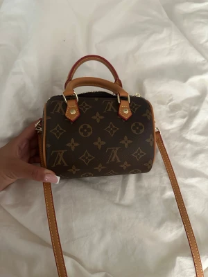 Brun monogram handväska mini - Snygg liten handväska i brunt med klassiskt monogrammönster och detaljer i beige och guld. Väskan har både handtag och en lång axelrem, perfekt för att bära crossbody. Tillverkad i tåligt konstläder med guldfärgade metalldetaljer.