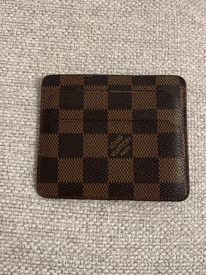 L och V brun rutig kortfodral - Säljer ett klassiskt kortfodral från L och V i brunt och mörkbrunt med det ikoniska rutiga Damier-mönstret. Tillverkat i tåligt skinn med flera kortfack. Perfekt för dig som vill ha en stilren och lyxig accessoar i fickan.