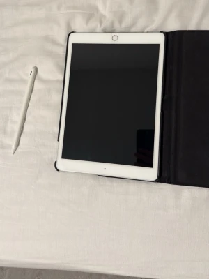 iPad med svart fodral och penna - Säljer en vit iPad med svart skyddsfodral och tillhörande vit styluspenna. Fodralet har ett runt fönster som visar Apple-loggan på baksidan. Perfekt för studier, anteckningar eller kreativt arbete.