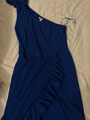 Blå oneshoulder klänning med volang - Snygg blå oneshoulder klänning med volangdetalj längs framsidan. Klänningen har en asymmetrisk axel och är tillverkad i ett mjukt, lätt material som ger ett fint fall. Perfekt för dig som vill sticka ut med en trendig och elegant look.