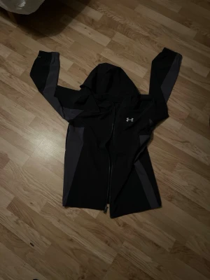 Svart vindjacka med huva Under Armour - Snygg svart vindjacka från Under Armour med huva och dragkedja framtill. Jackan har mörkgrå detaljer längs ärmar och sidor samt en vit logga på bröstet. Perfekt för blåsiga dagar och sportiga outfits.