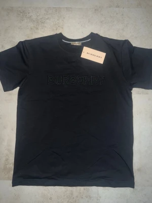 Svart Burberry t-shirt med broderad logga - Snygg svart t-shirt från Burberry med broderad logga framtill. Klassisk passform och rund halsringning. Tillverkad i mjuk bomull som känns skön mot huden. Perfekt för dig som gillar stilrena och exklusiva plagg. Storlek är M