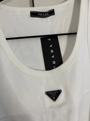 Vit linnetopp från Prada - Snygg vit linnetopp från Prada med rund halsringning och ikonisk metallogo framtill. Tillverkad i mjuk bomull som känns skön mot huden. Perfekt att styla med jeans eller kjol för en clean och trendig look.