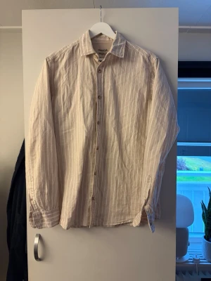 Linneskjorta Jack & Jones - Säljer en beige skjorta med vita vertikala ränder. Skjortan har klassisk krage, knäppning framtill och långa ärmar med knapp vid manschetten. Tillverkad i mjuk bomull, perfekt för en avslappnad men stilren look. Strl S passar M