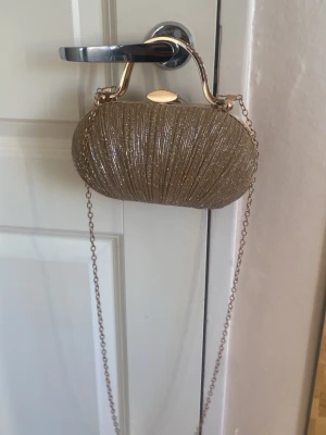 Glittrig guldig kuvertväska med kedja - Säljer en glittrig kuvertväska i guld med hårt skal och snygg metallkedja. Väskan har ett elegant handtag upptill och stängs med ett metallspänne. Perfekt accessoar för att lyfta din outfit med lite extra bling.