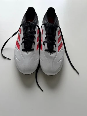 Adidas fotbollsskor  - Snygga fotbollsskor från Adidas i vitt med röda och svarta detaljer. Skorna har klassiska tre ränder på sidan, svart snörning och svart insida. Ovandelen är i syntetmaterial och designen är sportig och modern, perfekt för dig som vill sticka ut på planen.