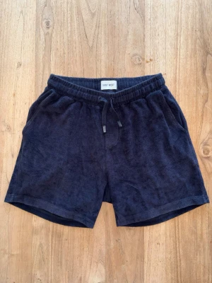 Mörkblå frottéshorts från East West - Mörkblå shorts i mjuk frotté från East West. Modellen har resår i midjan med snörning och två sidofickor. Perfekta för sommardagar eller att chilla i hemma. Enkel och stilren design med avslappnad passform.