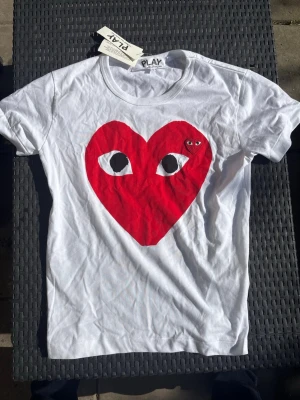 Vit t-shirt med rött hjärta - Säljer en vit t-shirt med ett stort rött hjärta med ögon på bröstet. T-shirten har rund hals och korta ärmar. Perfekt för dig som gillar grafiska tryck och vill sticka ut lite extra.