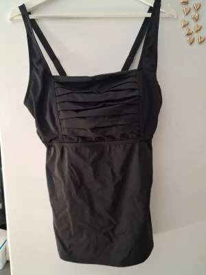 Svart tankini med veckad front - Stilren svart tankini med breda axelband och snygga veckade detaljer över bysten. Klassisk modell med markerad midja och justerbara axelband. Materialet ser ut att vara stretchigt och bekvämt, perfekt för stranden eller poolen.