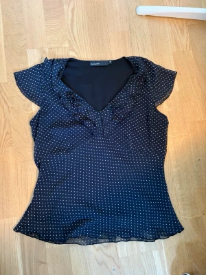 Svart prickig blus med volang - Snygg svart blus med vita små prickar och volangdetaljer vid v-ringningen. Blusen har korta ärmar och är tillverkad i ett tunt, lätt material som ger ett luftigt intryck. Perfekt att styla med jeans eller kjol för en trendig look.
