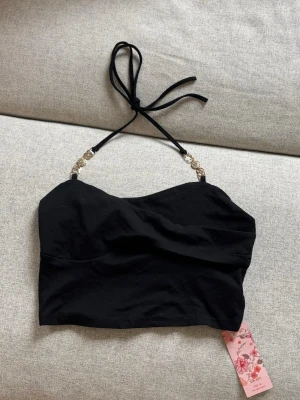 Svart bandeau topp med gulddetaljer - Snygg svart topp med tunna axelband som knyts i nacken och guldfärgade detaljer vid banden. Toppen är croppad och har en enkel, stilren design som passar perfekt till många olika looks. Nyskick med lappen kvar. Från shein.
