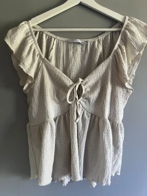 Lager 157 Beige blus med volang och knyt storlek M  - Supersöt vit blus med volangdetaljer på ärmarna och knyt framtill. Tillverkad i ett lätt, skrynkligt tyg som ger en avslappnad vibe. Perfekt till jeans eller kjol för en chill look.
