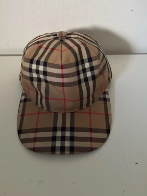 Beige rutig keps från Burberry - Säljer en klassisk beige keps från Burberry med det ikoniska rutmönstret i svart, vitt och rött. Kepsen har justerbar snapback i svart och en patch med texten 'BURBERRY LONDON ENGLAND' bak. Perfekt accessoar för en trendig och stilren look.