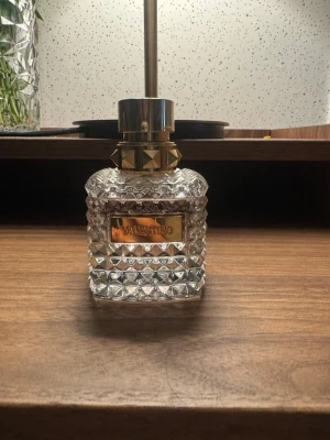 Valentino parfym med nitar och glas - Säljer en lyxig parfym från Valentino i en transparent glasflaska med coolt nitat mönster och guldfärgad etikett. Flaskan har en unik design med gulddetaljer och en elegant, fyrkantig form. Perfekt för dig som gillar exklusiva dofter och snygg inredning. Den är använd några gånger som man ser men väligt mycket kvar