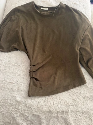 Olivgrön långärmad assymmetrical topp - Snygg olivgrön långärmad tröja i mjuk sammetsliknande material. Tröjan har rund halsringning och en lätt figursydd passform med diskreta veck i sidan. Perfekt för en trendig och avslappnad look.