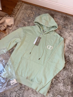 Mintgrön hoodie med broderad logga - Snygg mintgrön hoodie med huva och dragsnören. På bröstet finns en broderad vit logga med bokstäverna 'CD'. Hoodien har en klassisk känguruficka framtill och ribbade muddar vid ärmslut och nederkant. Perfekt för en chill och trendig look. Unik och jätte snygg perfekt för att sticka ut lite passar även M 