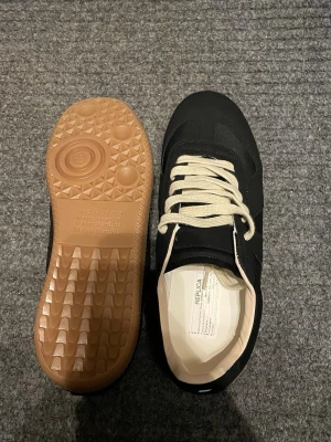 Svarta sneakers med beige sula - Stilrena svarta sneakers med beige snörning och kontrasterande ljusbrun gummisula. Ovandelen är i mocka och textil, med klassisk låg silhuett och retrovibbar. Perfekta för dig som gillar minimalistisk och tidlös design.