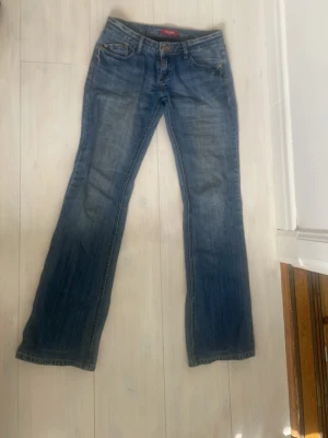 Blå bootcut jeans med låg midja - Säljer ett par klassiska blå bootcut jeans med låg midja och snygga slitningar. Jeansen har fem fickor och stängs med dragkedja och knapp. Perfekta för en avslappnad och trendig look. Innerbenslängd 83 midja 37
