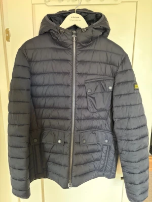 Mörkgrå Barbour dunjacka med huva - Snygg mörkgrå Barbour dunjacka med huva och dragkedja framtill. Jackan har tre fickor med lock och tryckknappar samt en patch på ena ärmen. Quiltad design och justerbar huva med snörning. Perfekt för kalla vinterdagar.