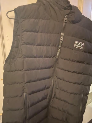 Svart dunväst från EA7 Emporio Armani - Snygg svart dunväst från EA7 Emporio Armani med quiltad design och dragkedja framtill. Västen har hög krage, två fickor med dragkedja och logga på bröstet. Perfekt för lager-på-lager och streetstyle. Köpt på mall of scandinava 