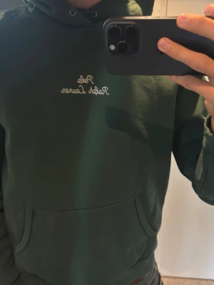 Mörkgrön hoodie från Polo Ralph Lauren - Mörkgrön hoodie från Polo Ralph Lauren med broderad logga på bröstet och liten vit häst på ärmen. Klassisk känguruficka framtill och justerbar huva med dragsko. Perfekt för en avslappnad och stilren look.