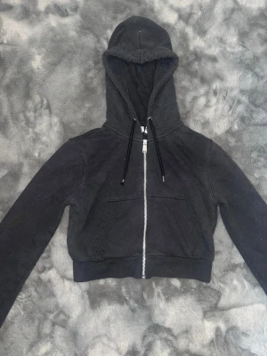 Svart croppad hoodie med dragkedja - Säljer en svart croppad hoodie med dragkedja framtill och två fickor. Hoodien har långa ärmar, ribbade muddar och justerbar huva med svarta snören. 