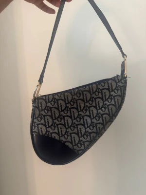 Blå axelväska i monogramtyg - Säljer en snygg axelväska i blått och beige monogramtyg med D-detaljer i metall. Väskan har en unik sadelform och justerbart axelband i läder. Perfekt för dig som gillar statement-accessoarer och vill ha något trendigt och klassiskt på samma gång. (Se bild kedjan funkar men själva hänget på kedjan är av) 