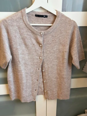 Beige kortärmad kofta från 157 - Söt beige kofta från 157 med korta ärmar och pärlknappar framtill. Koftan är stickad i ett mjukt material och har rund halsringning samt ribbade kanter. Perfekt att slänga över en topp för en chill look. Katt finns hemma 