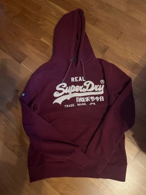 Vinröd hoodie från Superdry - Snygg vinröd hoodie från Superdry med stor vit logga och japansk text på bröstet. Klassisk känguruficka framtill, justerbar huva med dragsko och ribbade muddar. Tillverkad i mjukt bomullsmaterial som är skönt mot huden. Hoodien är i väldigt bra skick och storleken är M. Mitt pris är 399kr men det går att diskuteras.