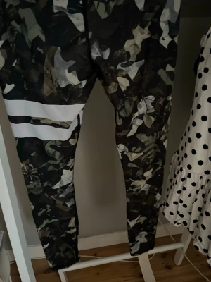 Camo leggings med vita ränder - Snygga leggings med camouflage-mönster i gröna, bruna och svarta toner samt två breda vita ränder på ena benet. Tight passform och stretchigt material som passar perfekt till en sportig eller avslappnad stil.